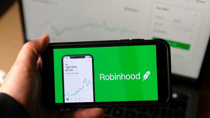 An iPhone is held up to the camera with a green Robinhood ad taking up the entire screen. Behind, a laptop displays stocks chart