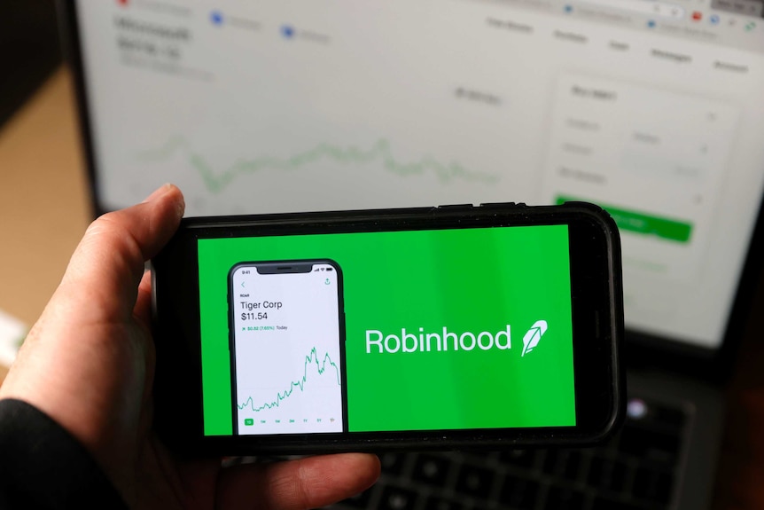 An iPhone is held up to the camera with a green Robinhood ad taking up the entire screen. Behind, a laptop displays stocks chart