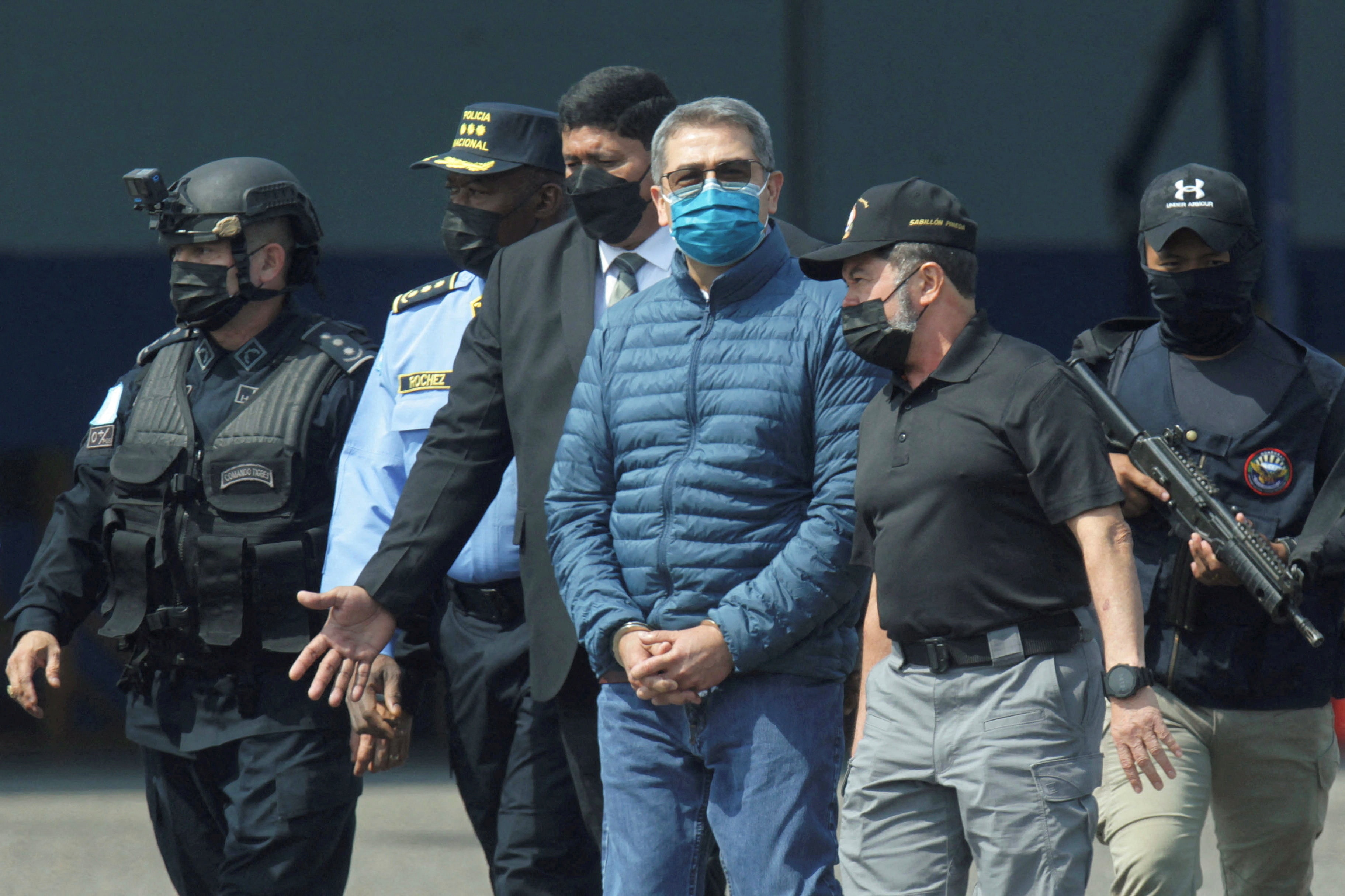 Juan Hernandez, wearing a medical face mask and puffer jacket, is surrounded by law enforcement officers.