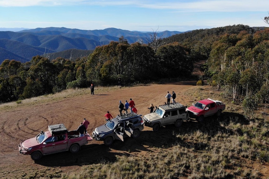 Aerial view of four trucks parked in a field with people standing next to them and mountains in the background.
