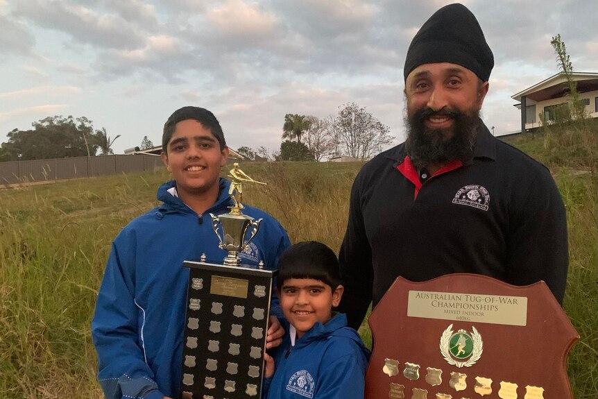 A man and two boys smile at the camera while they stand outside holding trophies and shields.