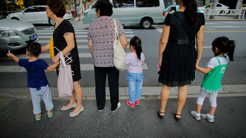 Three older women each hold the hand of a child at a pedestrian crossing