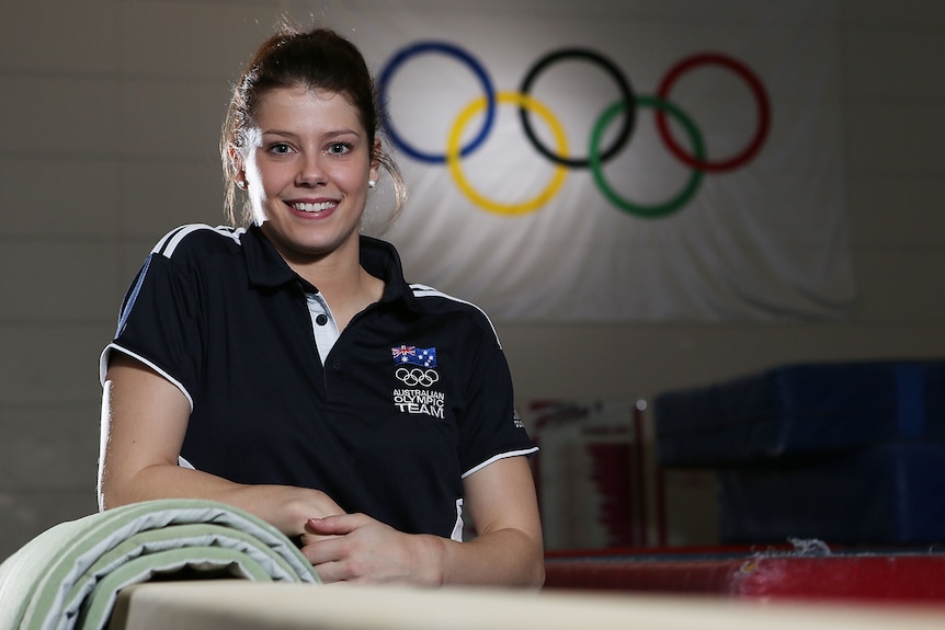 Gymnast Georgia Bonora leans on a balance beam while smiling for the camera in front of the Olympic rings.