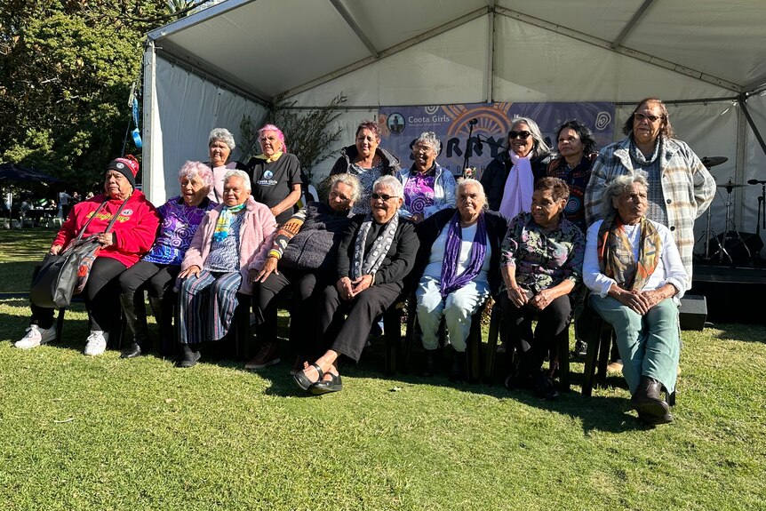 A group of seated older Aboriginal women pose for a photo together outside a marquee in a green park.