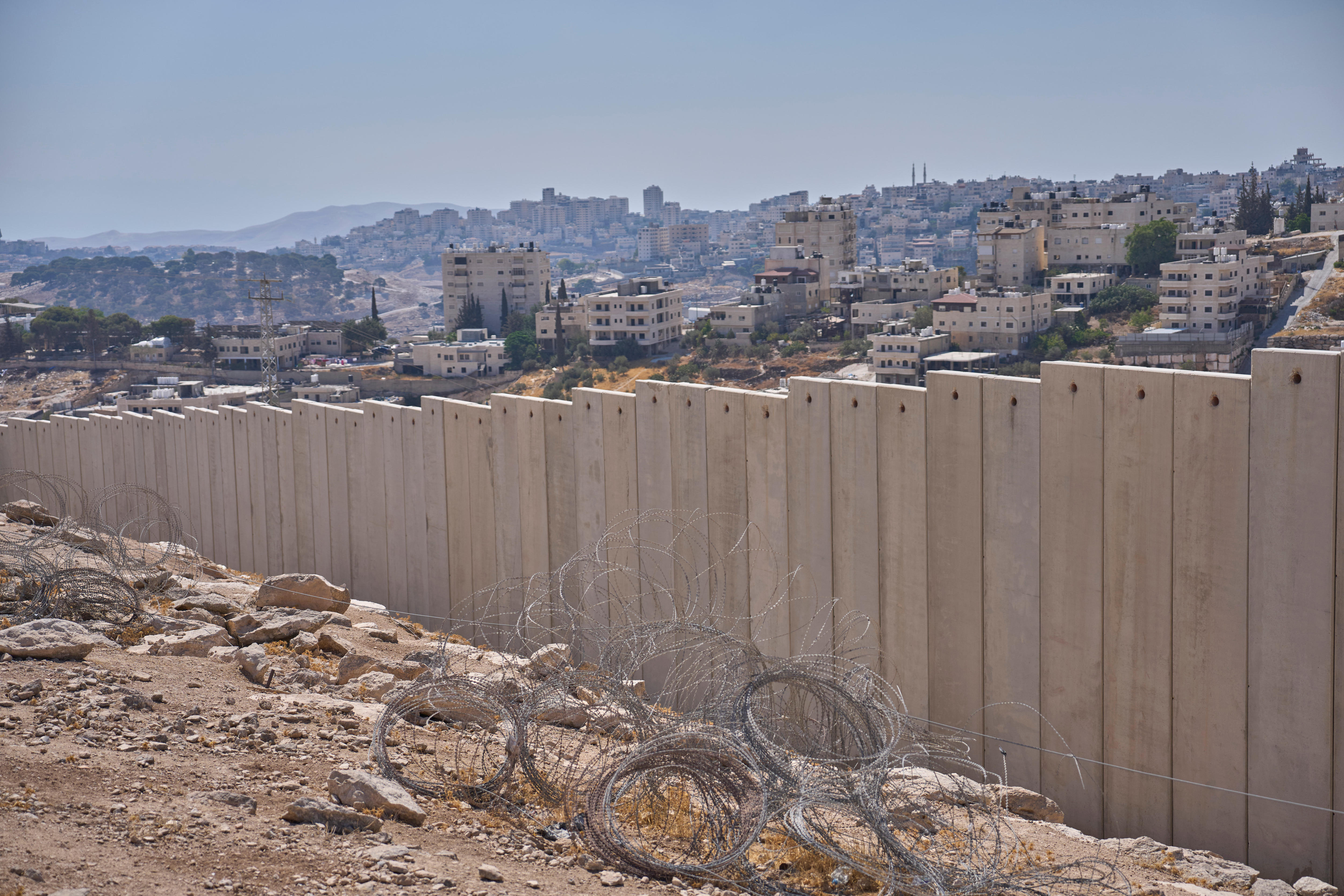 A large concrete barrier, made up of tall, lined-up slabs, runs in front of a collection of buildings on a dusty hillside.