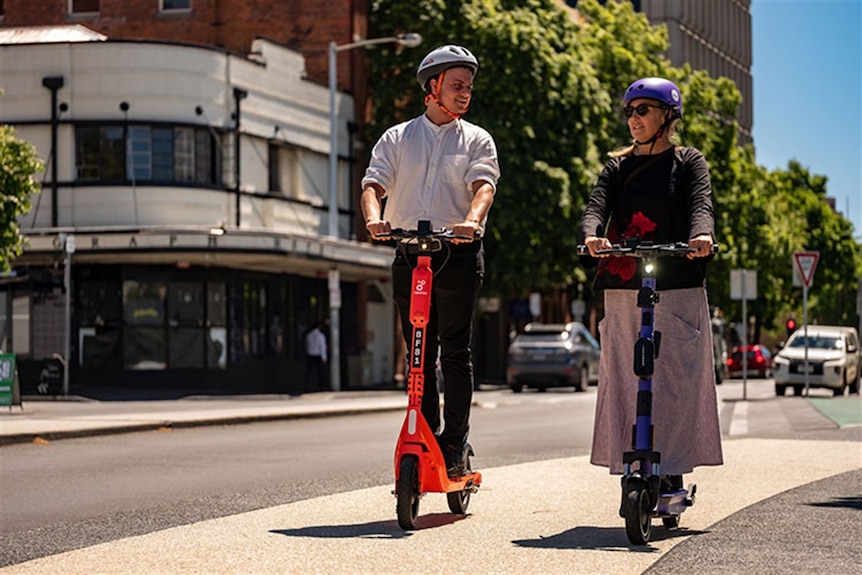 Male and female riding e-scooters on city footpath.