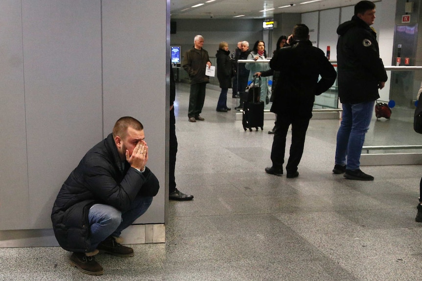 A man stands bent down with his head in his hands in shock. People mill about to the right.