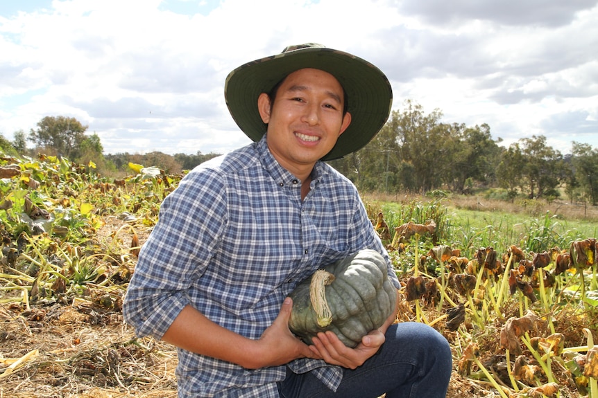 A man holding a pumpkin kneels in a vegetable patch and smiles at the camera.