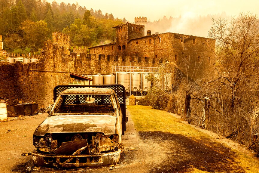 You view the charred remains of a ute parked in front the shell of a medieval-style building set against a smoky sky.