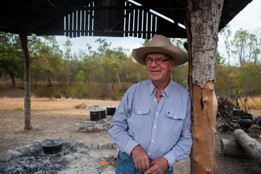 A man wearing an akubra leans against a tree-pole and looks to the side happily. 