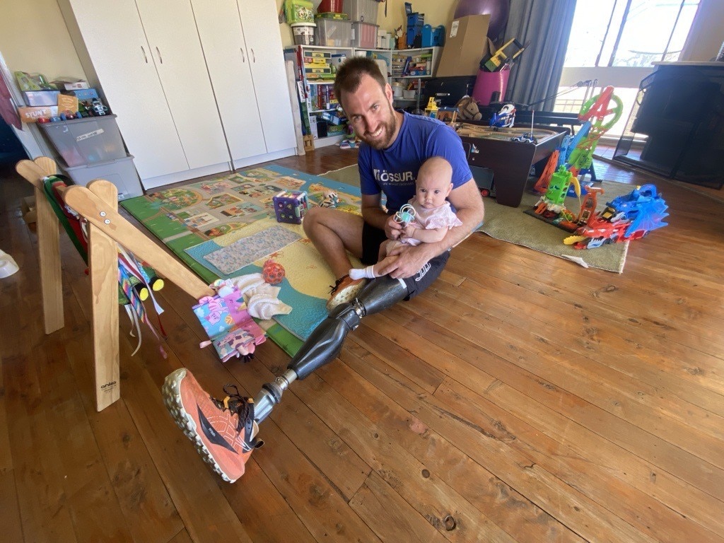 Aaron sorrindo para a câmera sentado no chão, segurando um de seus filhos pequenos