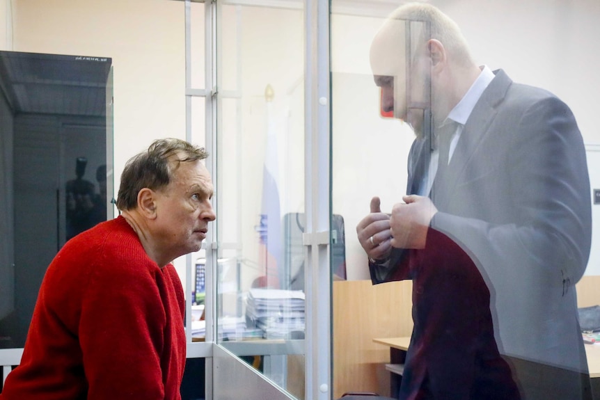 Sokolov sits behind a glass screen in courtroom in a red jumper. A man in a suit talks to him.