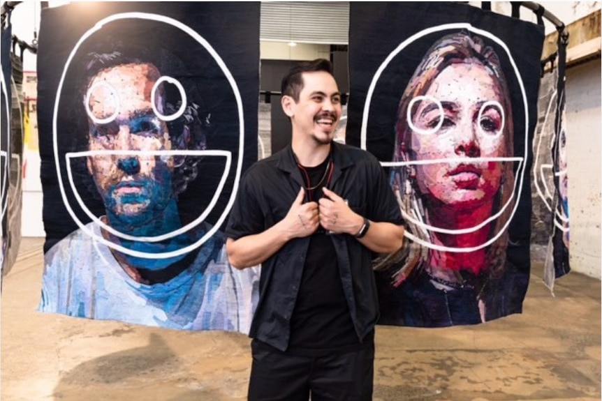 Artist Abdul Abdullah stands in a gallery in front of two large portrait tapestries
