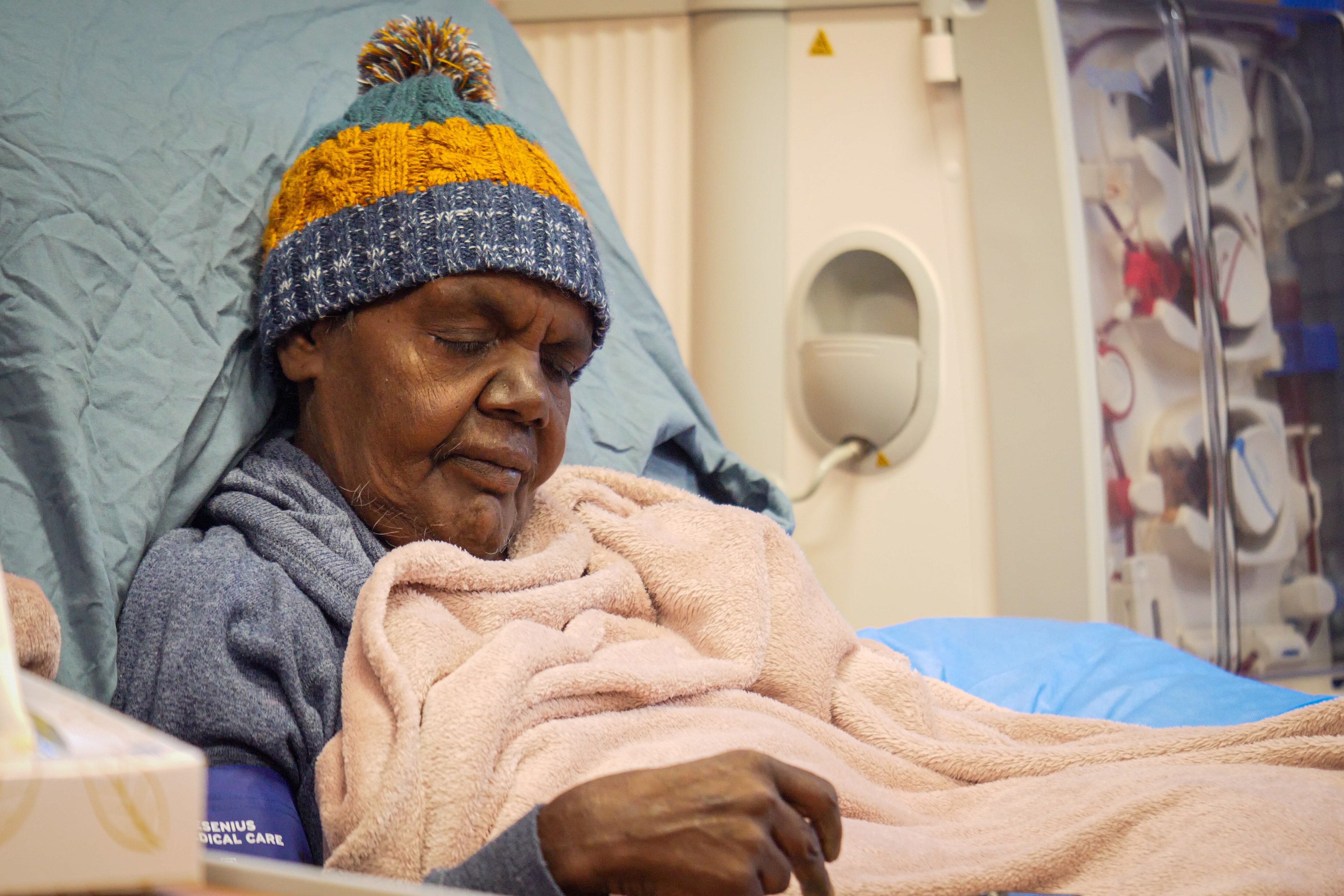 An older Aboriginal woman sits upright with her eyes shut in hospital bed. The dials of a dialysis unit are beside her.