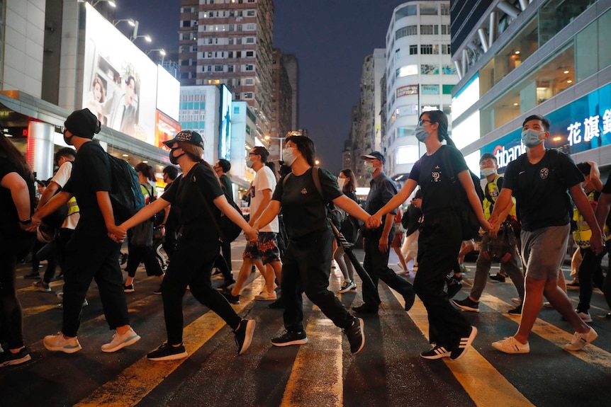 Young people, wearing black and with masks over their faces, hold hands while walking across the street.