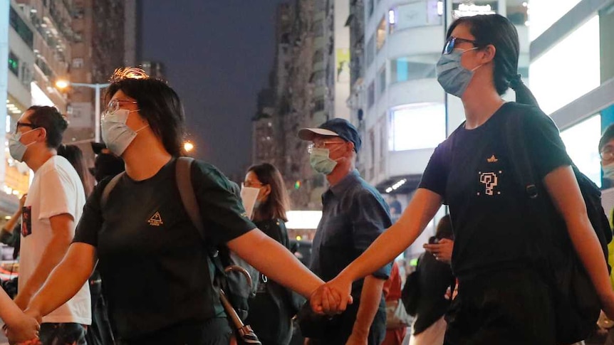 Young people, wearing black and with masks over their faces, hold hands while walking across the street.