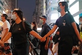 Young people, wearing black and with masks over their faces, hold hands while walking across the street.