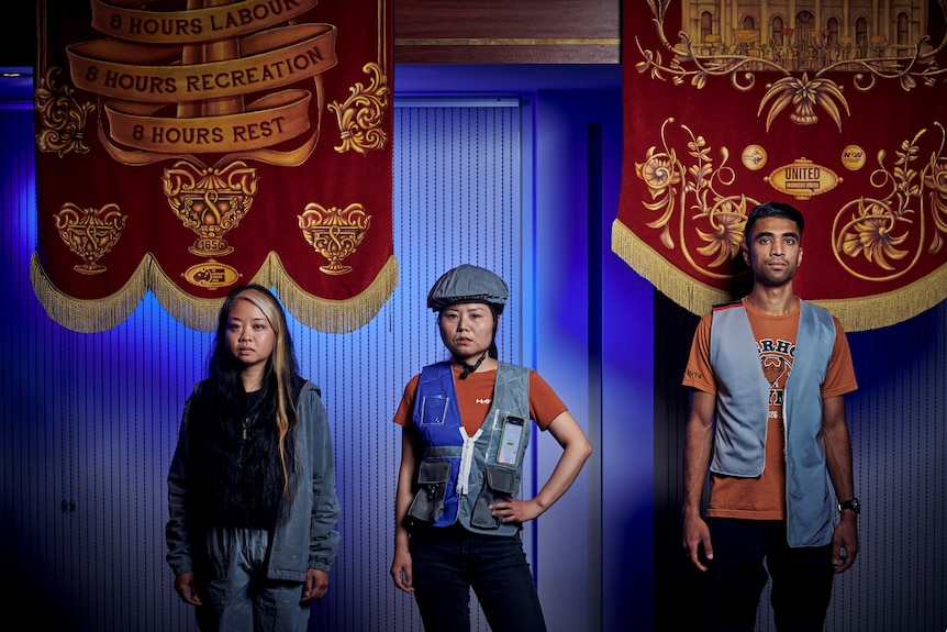 Three gig economy workers stand in front of red banners in Melbourne's Trades Hall, they are all performers in an artwork