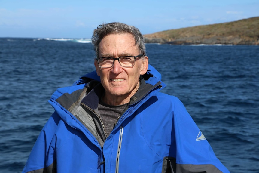 A man in a blue parka stands on a boat with ocean around him and land in the distance on the right.