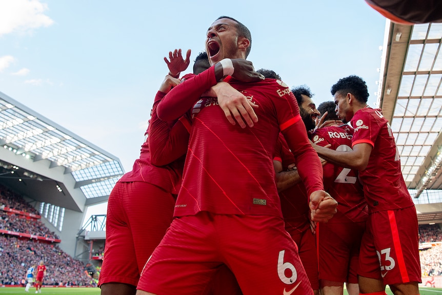 A Liverpool footballer stands roaring in triumph as his teammates hug and celebrate after a goal at Anfield. 