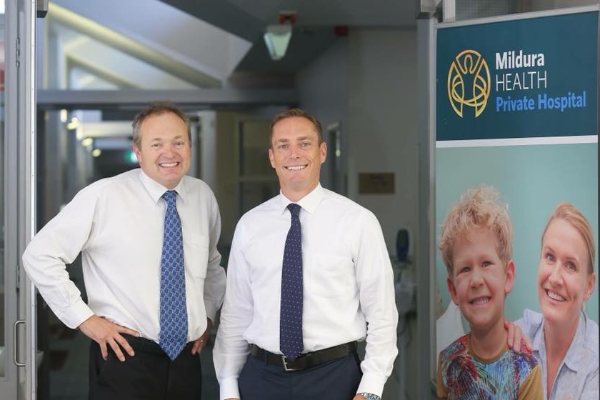 Two smiling men in business attire and sensible haircuts standing out the front of a private hospital.