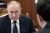 Russian President Vladimir Putin sits in a chair wearing a suit, looking at another man, whose back of his head is visible