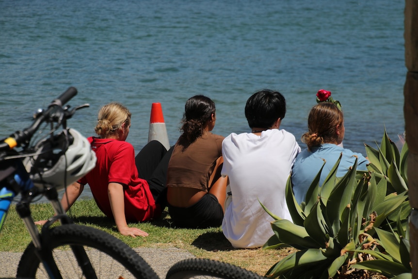 A group of young people sit and look at the river with their backs to the camera. 