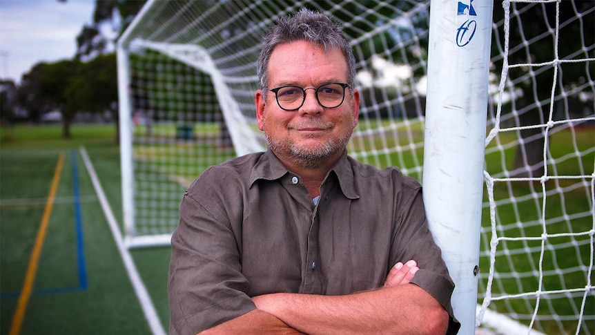 A man smiles, leaning against a soccer goal post.