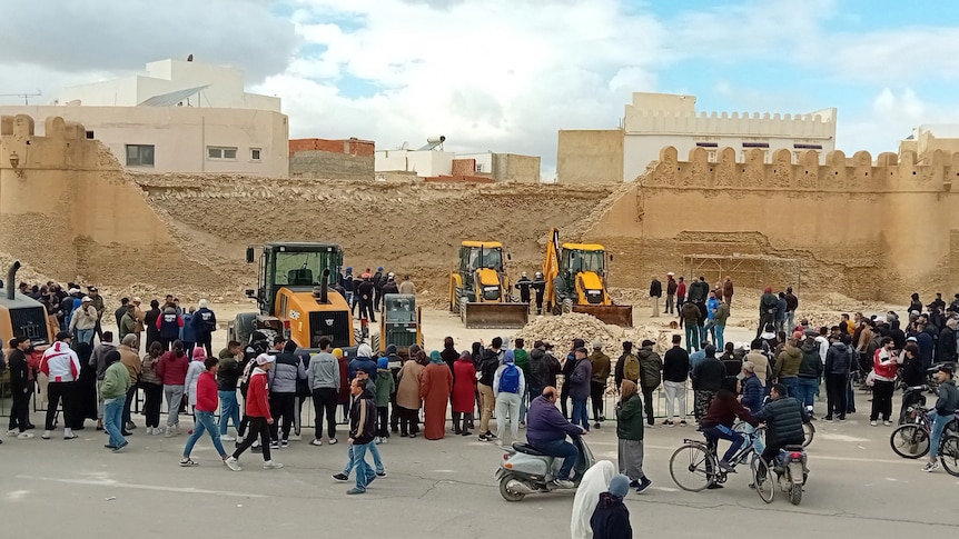 A crowd of people standing near a beige coloured wall with a collapsed section and yellow tractors in front.