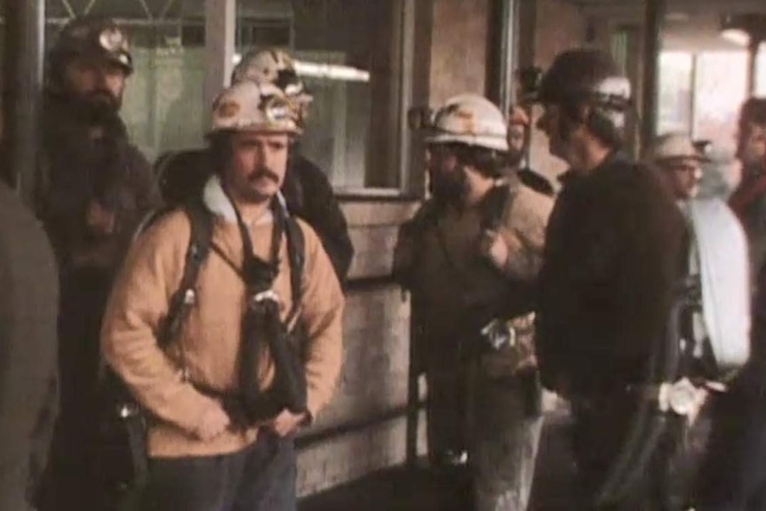 Grainy archival vision of ten mine rescuers standing in front of a brick building. All wear hard hats with head torches.