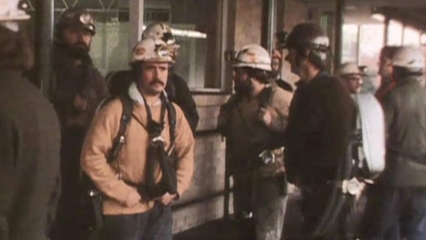 Grainy archival vision of ten mine rescuers standing in front of a brick building. All wear hard hats with head torches.