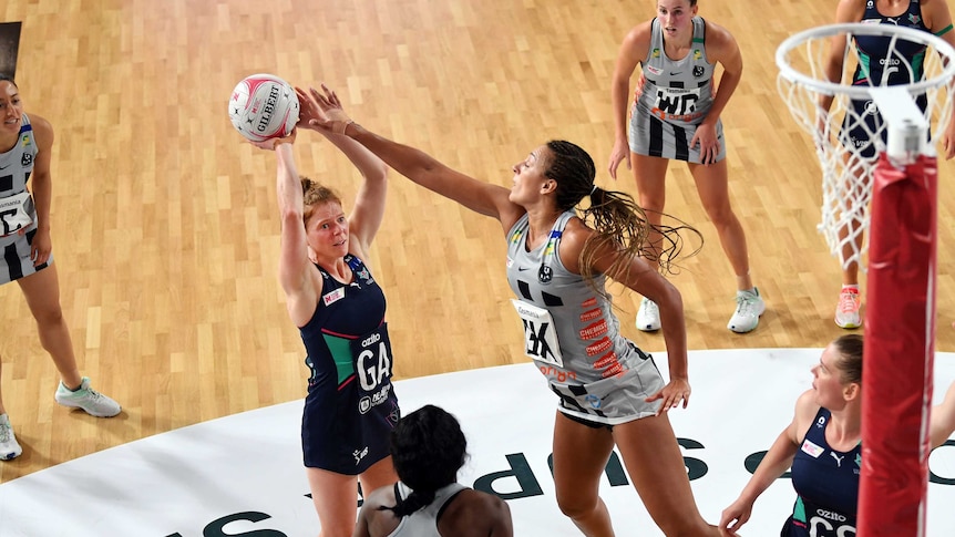 A netballer grimaces as she tries a two-point shot, while a defender stretches her arm out to block.