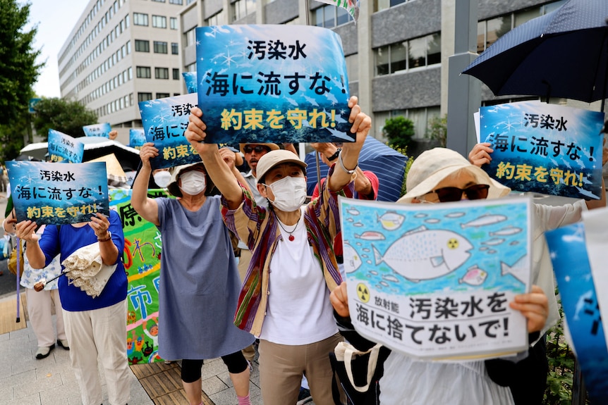 A group of middle-aged Japanese men and women hold up blue, ocean-themed posters on a street while wearing masks.
