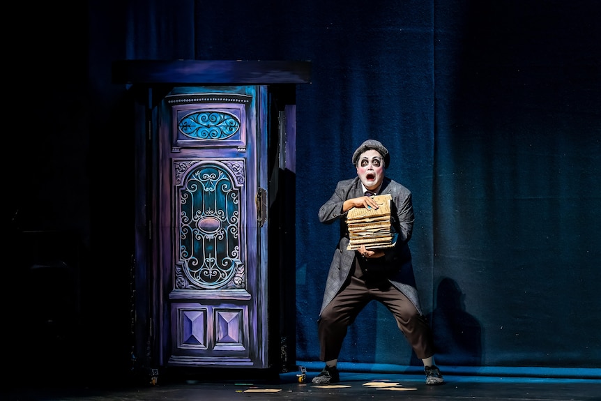 On stage, a man in a face of make-up has a surprised expression and is holding a stack of books. He's next to a wardrobe.