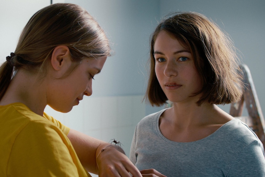 Two young women standing close by each other in perhaps a kitchen. One has a spider on her hand.