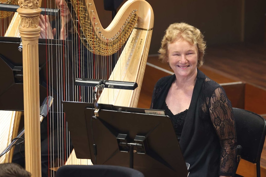 Harpist Jill Atkinson sitting in her place on the stage, next to her harp, smiling in her final performance before retiring.