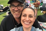 A couple smiling to the camera (selfie) sitting at an outdoor picnic.