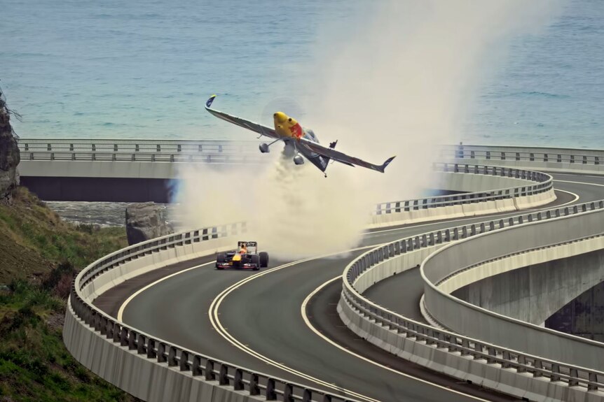 A plane flies low above a race car on a curved road bridge on the Great Ocean Road.