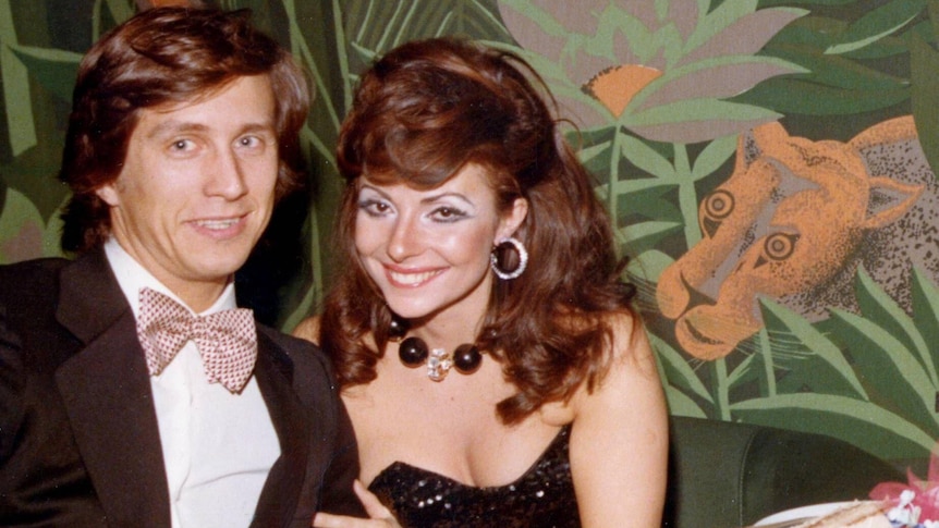 Maurizio Gucci, in tuxedo and pink bow tie, sits with Patrizia Reggiani, wearing a black ball gown, in front of floral wallpaper