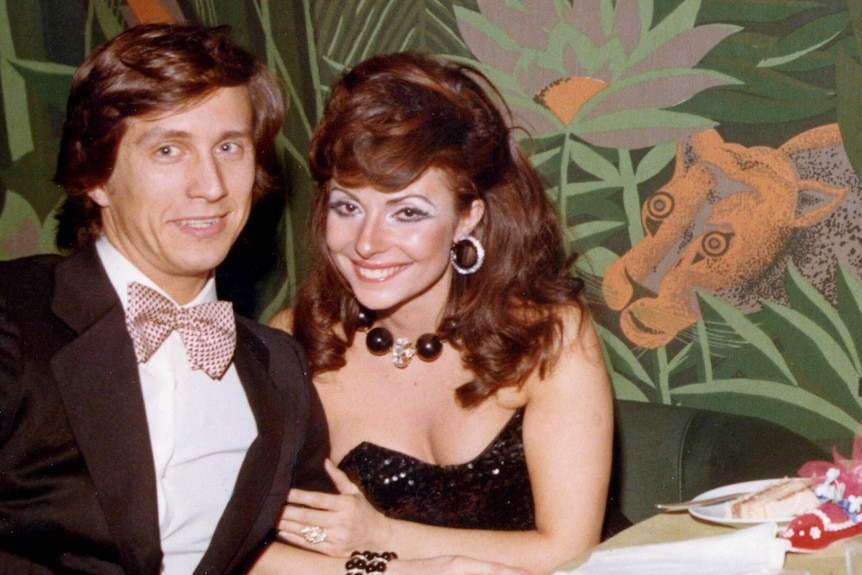 Maurizio Gucci, in tuxedo and pink bow tie, sits with Patrizia Reggiani, wearing a black ball gown, in front of floral wallpaper