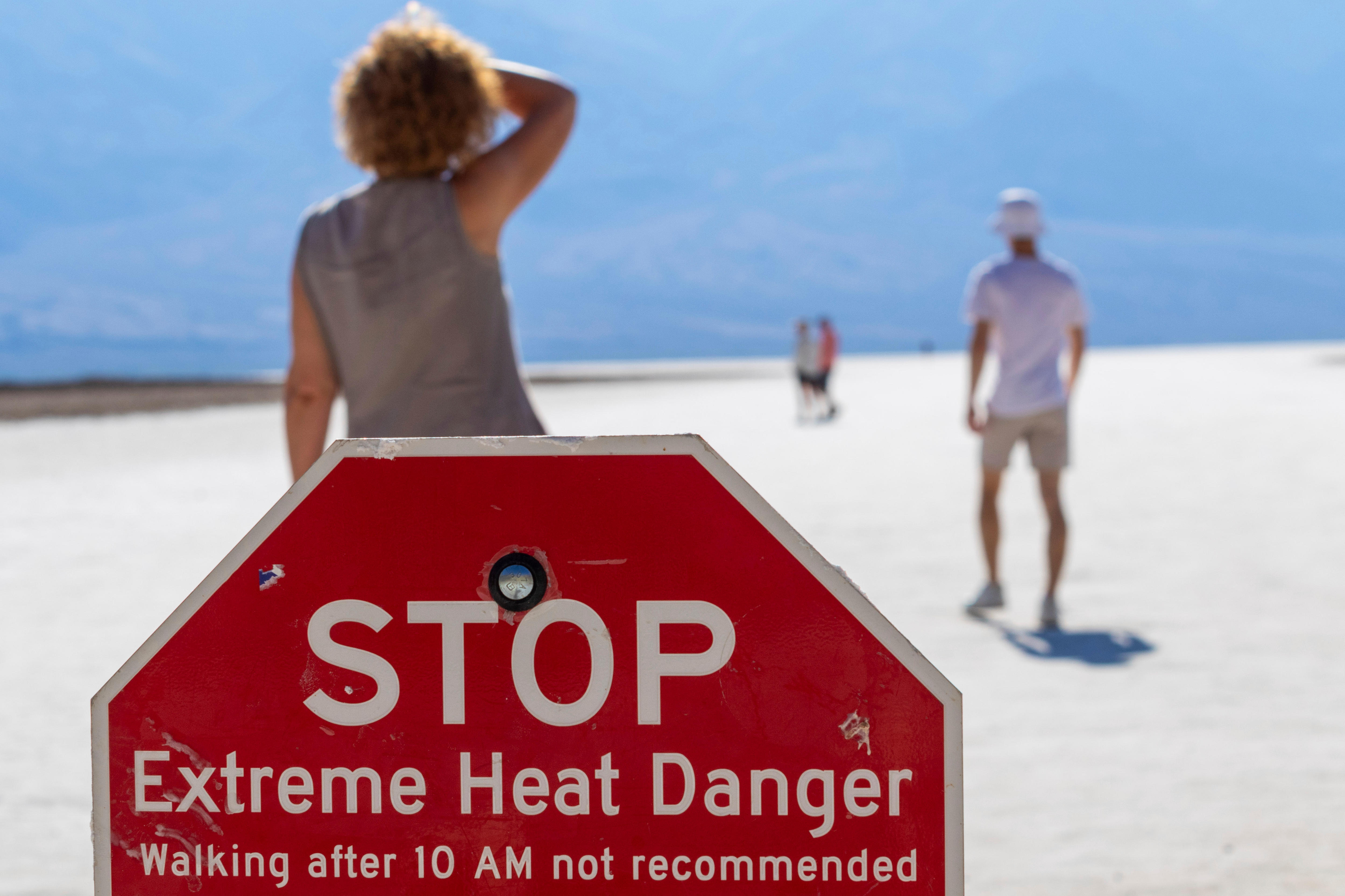 Four people walk across white sand in front of a red sign reading 'STOP: Extreme Heat Danger'.