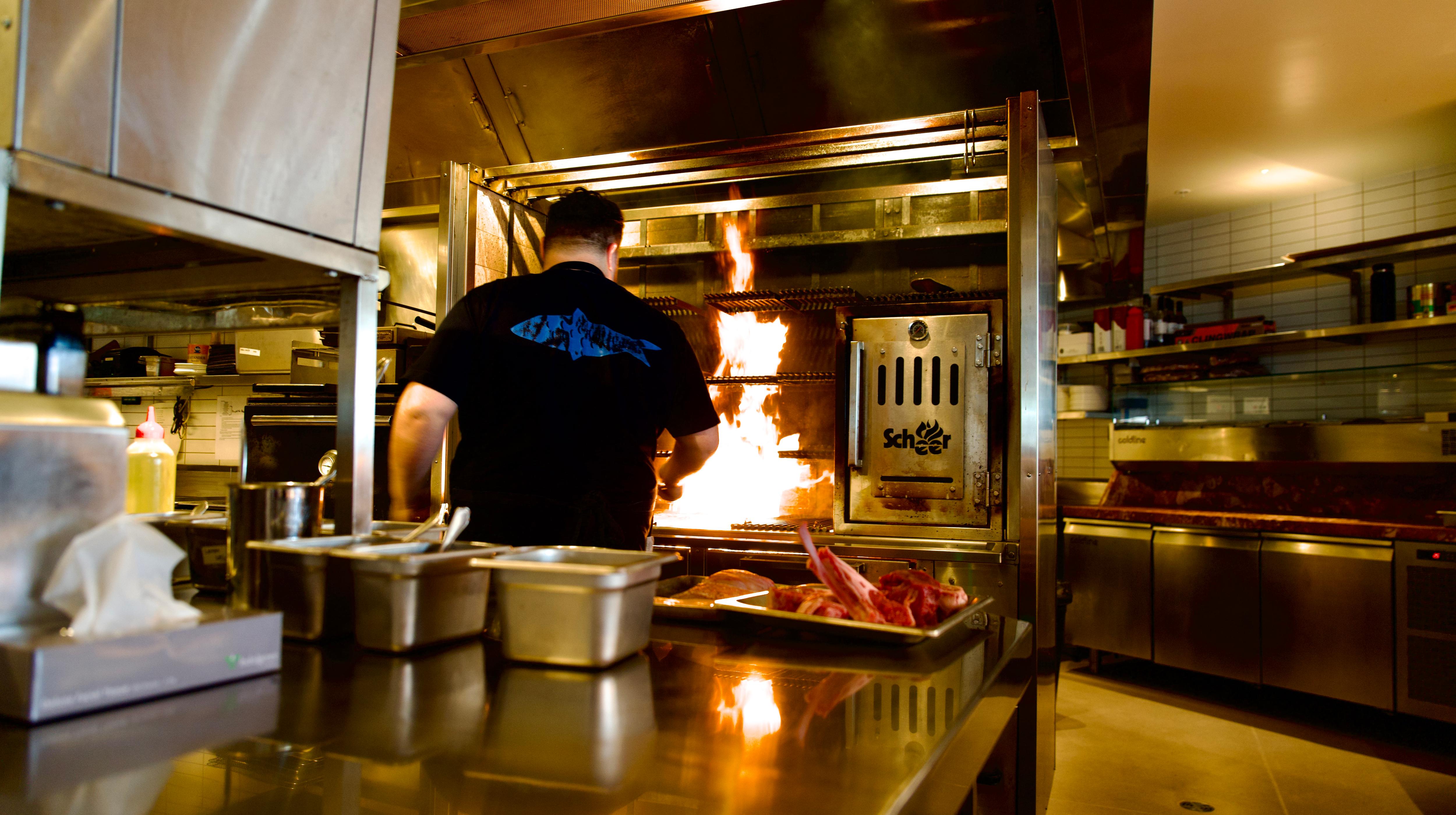 Head chef Luke Bourke stands in a commercial kitchen with his back to the camera, working over a flaming grill