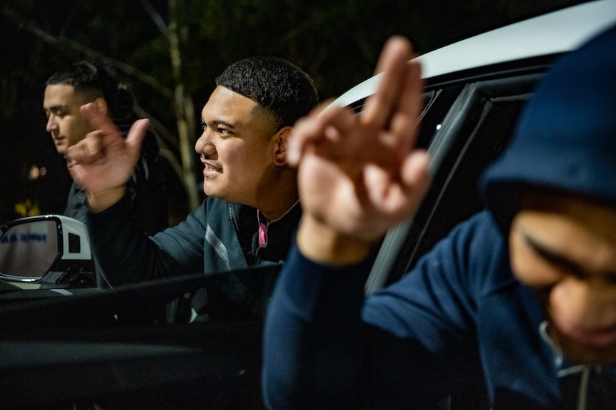 Two men shake their hands while pointing as they sit in the open doors of a car at night.