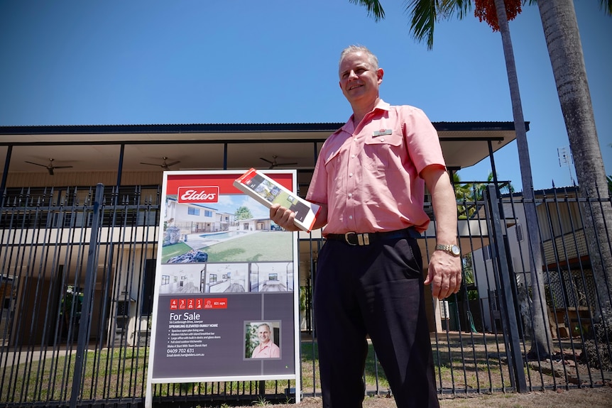 A man in a collared shirt and long trousers standing front of a house with a 'for sale' sign at the front, holding brochures.