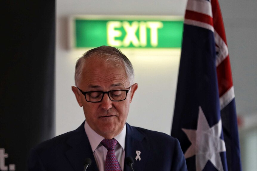 Malcolm Turnbull, wearing glasses, looks down towards a lectern. Behind him is a green EXIT sign and an Australian flag.
