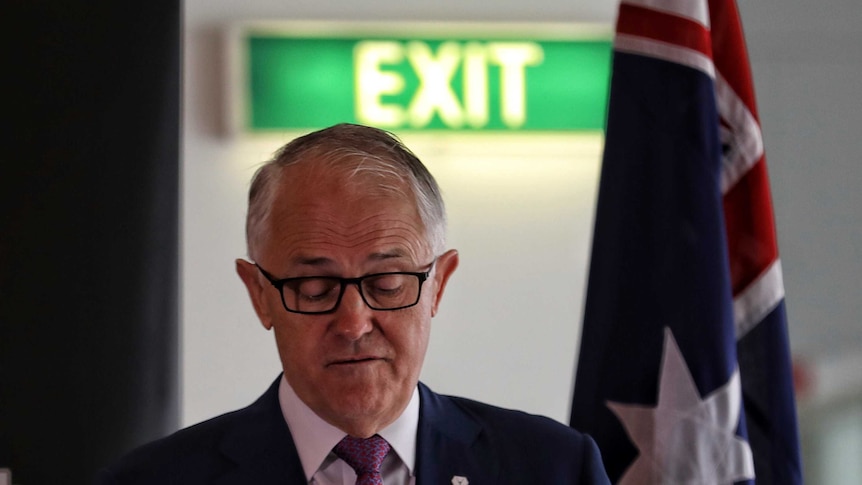 Malcolm Turnbull, wearing glasses, looks down towards a lectern. Behind him is a green EXIT sign and an Australian flag.