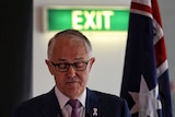 Malcolm Turnbull, wearing glasses, looks down towards a lectern. Behind him is a green EXIT sign and an Australian flag.