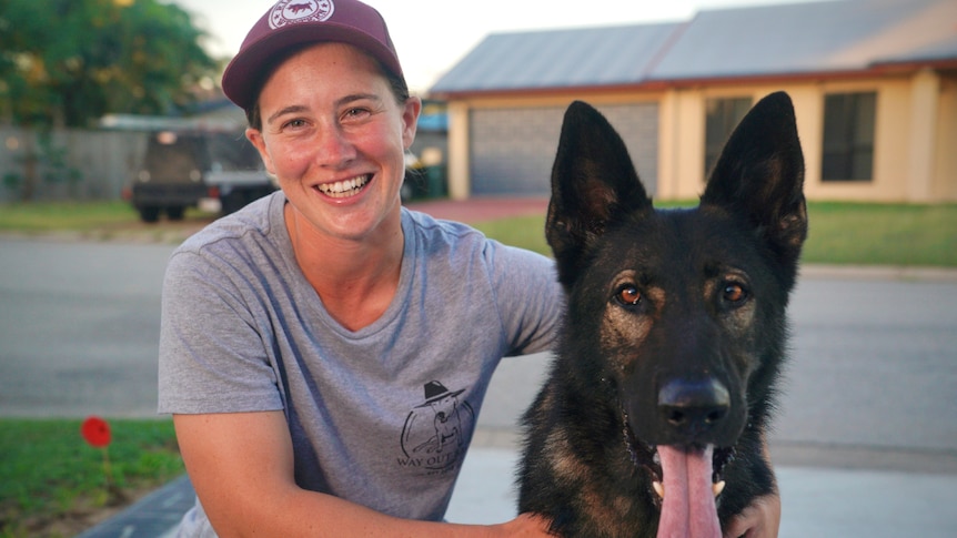 A woman squats beside a large black German shepherd-type dog.