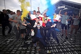 Leeds United fans hold blue and yellow flares and smile outside a building that says Leeds United Football Club Store