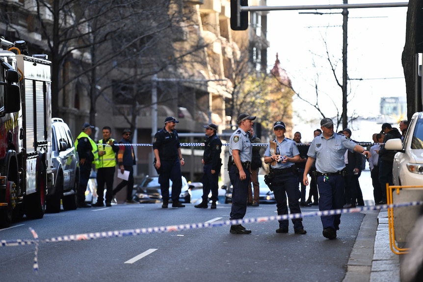 Police stand between emergency vehicles in a Sydney street, behind police tape.
